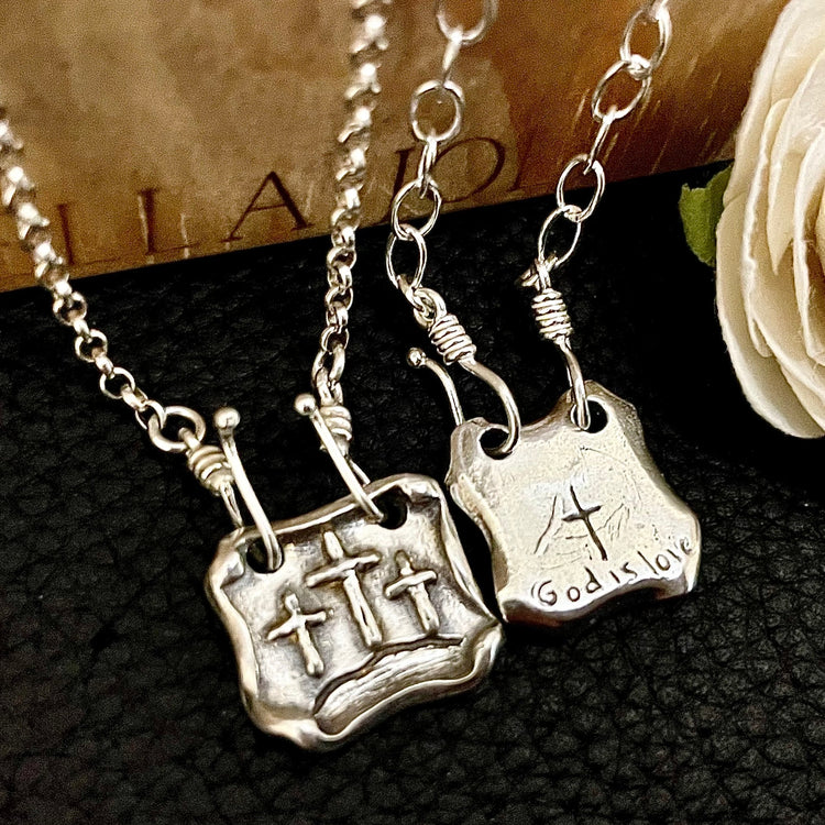 Two pendant necklaces with front side of three crosses of calvary and back side showing custom text of God is love.  Interchangeable pendants hanging from sterling silver chains laying on black leather and wood background. 