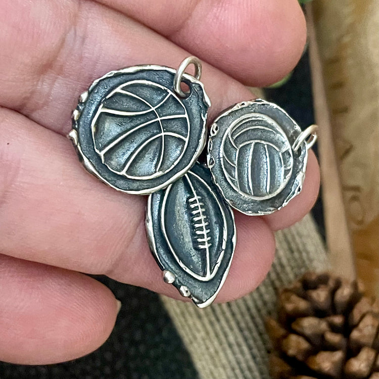 Three silver sports-themed pendants held in a hand with a blurred natural background.Can be personalized on backside. Sitting on a textured surface and pine cone in the background. Basketball, football and volleyball shown with baseball/softball and soccer also available.  