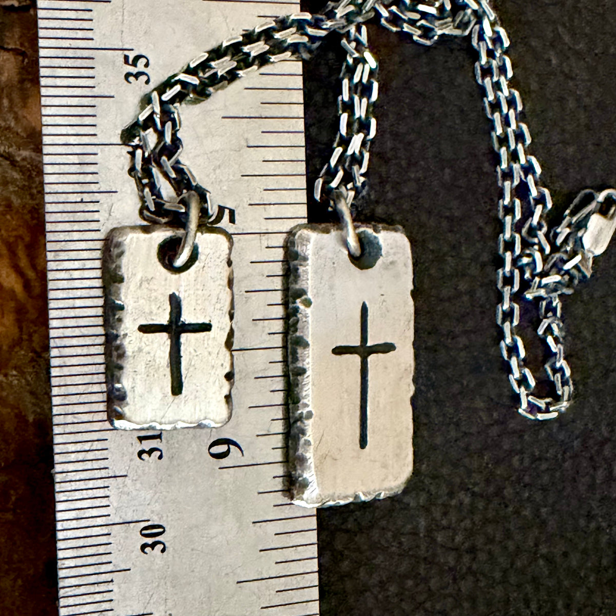 Two silver rectangular pendant necklaces with engraved crosses, shown next to a metal ruler for scale on a dark textured background.Both sizes with chains: Rustic fine silver cross necklaces for men in 3/4 inch and 1 1/4 inch sizes
