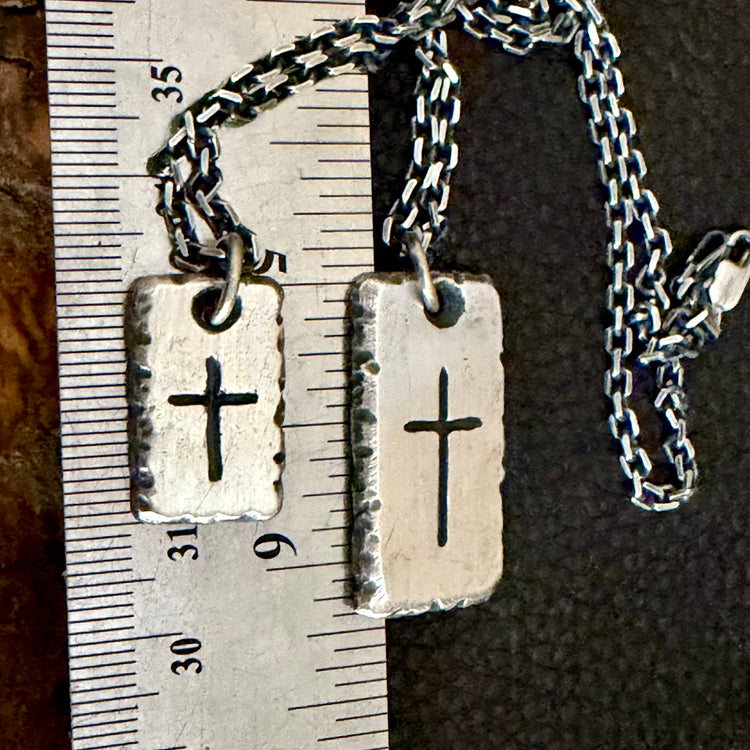 Two silver rectangular pendant necklaces with engraved crosses, shown next to a metal ruler for scale on a dark textured background.Both sizes with chains: Rustic fine silver cross necklaces for men in 3/4 inch and 1 1/4 inch sizes