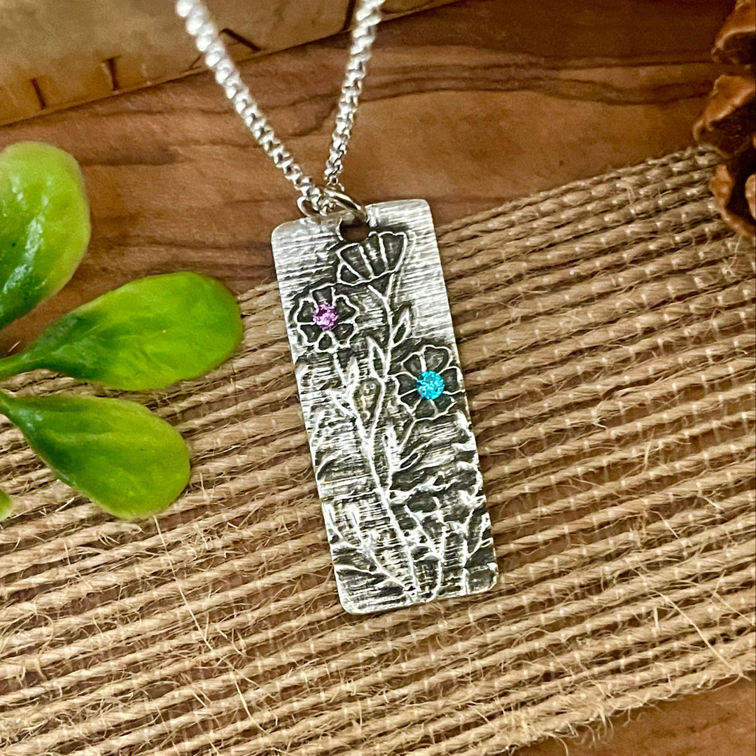 Sterling Silver necklace with a rectangular pendant featuring raised floral designs and crystal birthstone gemstones on a textured wooden surface.