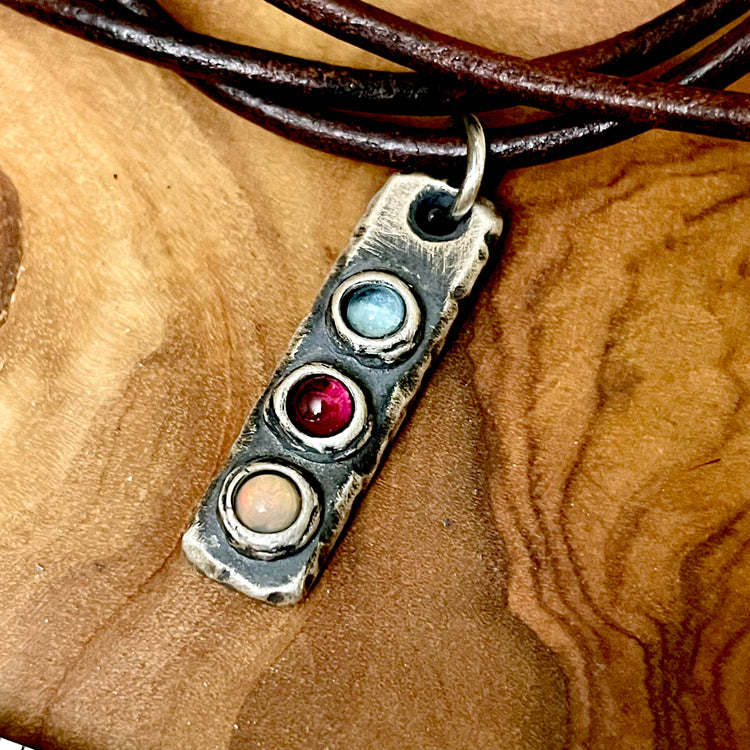 Rustic Sterling Silver Bar Necklace pendant featuring three custom colored birthstones on a wooden surface hanging from leather necklace. 