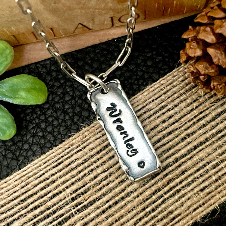Personalized Sterling Silver Name Necklace with custom engraving, handcrafted with raised edges and heart below name at bottom. Hanging from 925 sterling silver staple paperclip style chain on black and wood background. 
