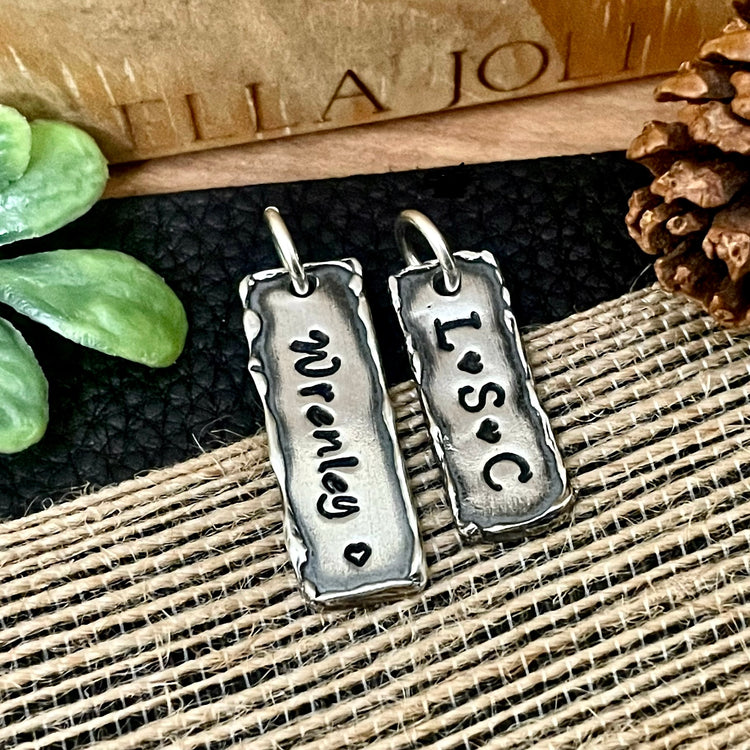 Two silver pendants with engraved text on a textured surface. One bar has a name with a heart and the other bar has three initials separated by hearts. Custom made your way. 