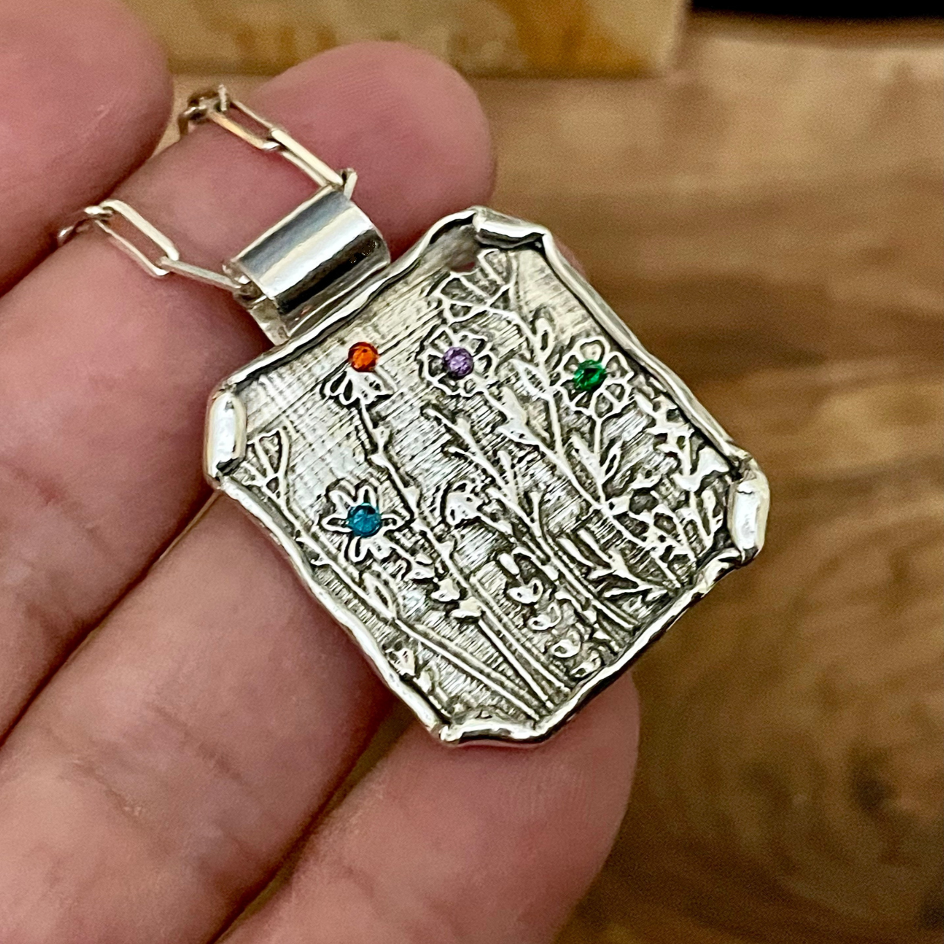 Silver pendant with intricate wildflower designs held in a hand against a wooden background