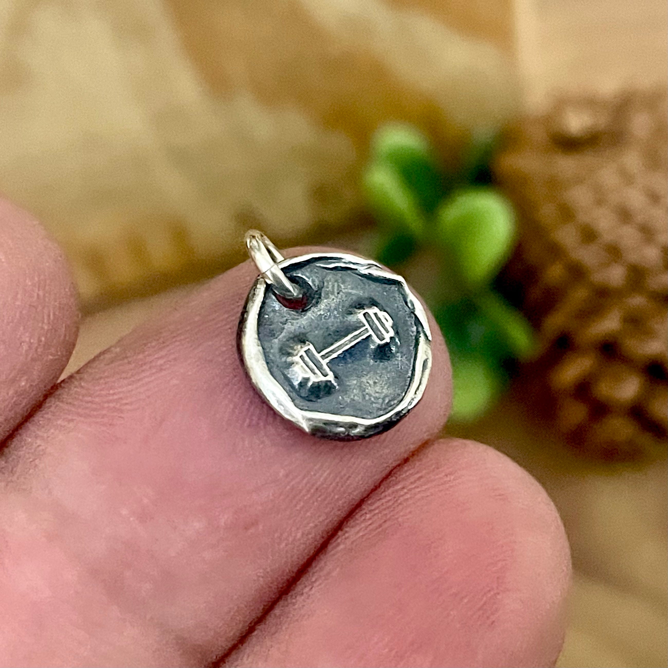 Silver fitness pendant with a barbell design held between fingers against a blurred natural background