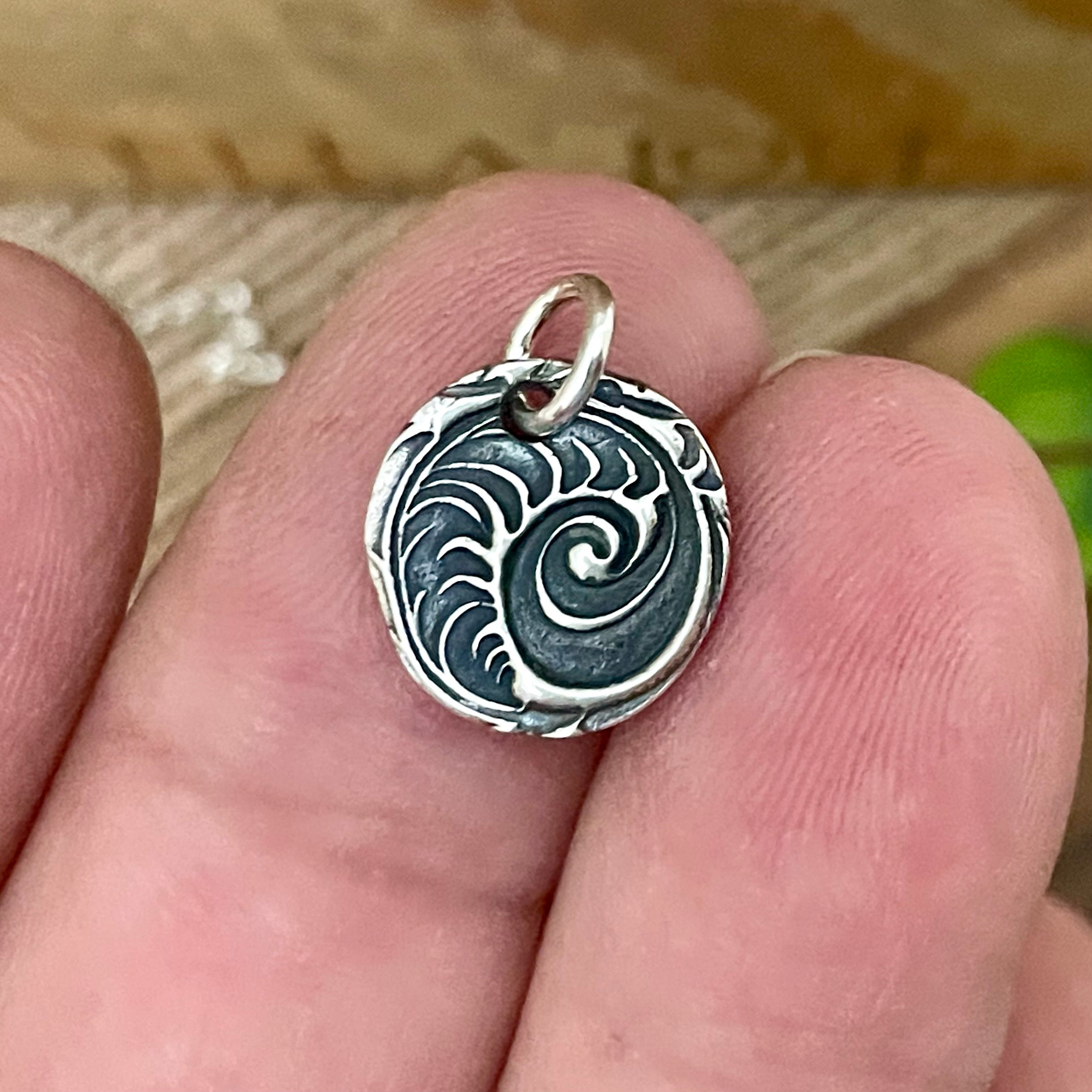 Silver pendant with intricate ocean wave design held between fingers against a blurred natural background