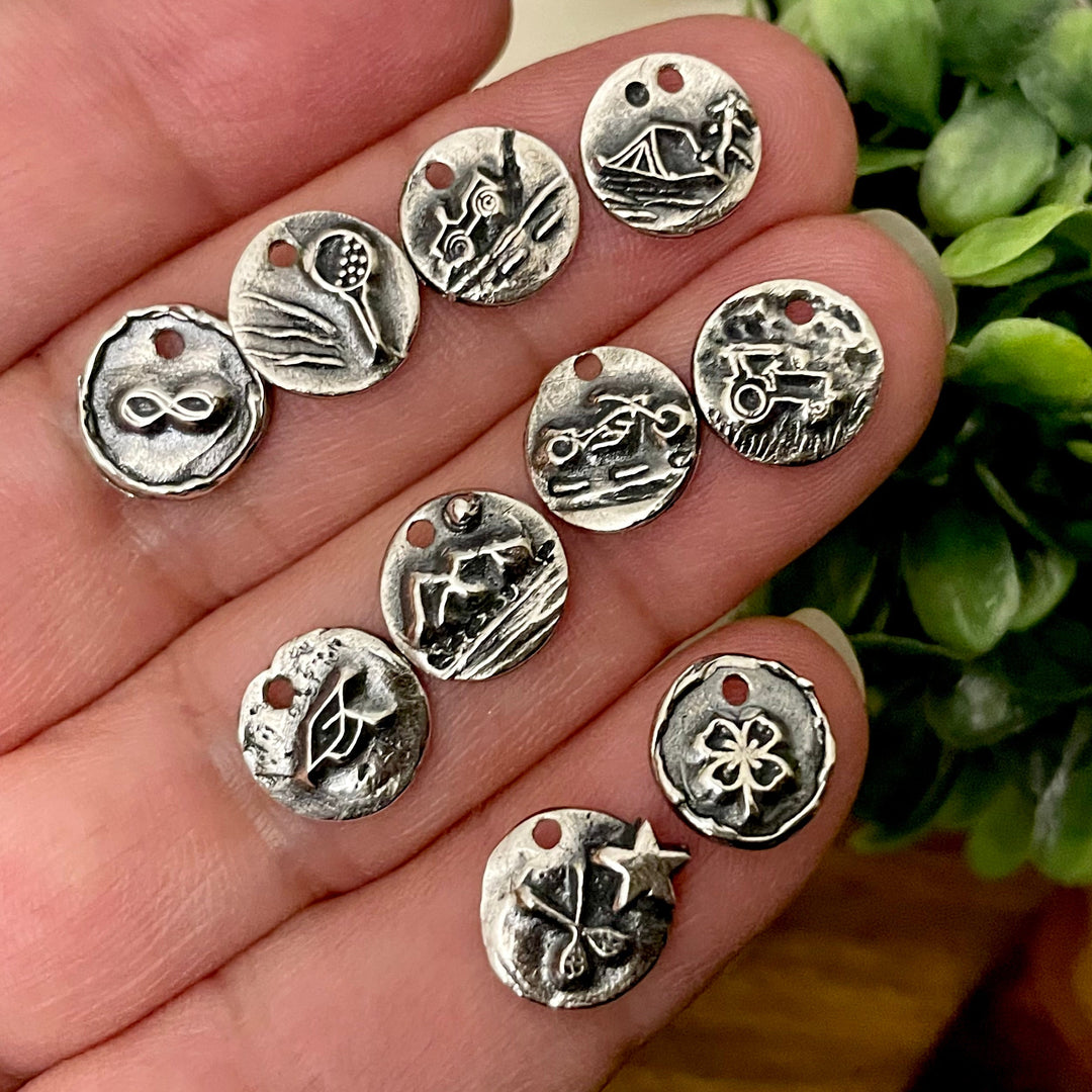 Sterling Silver charms with various designs held in a hand, with a green plant in the background.