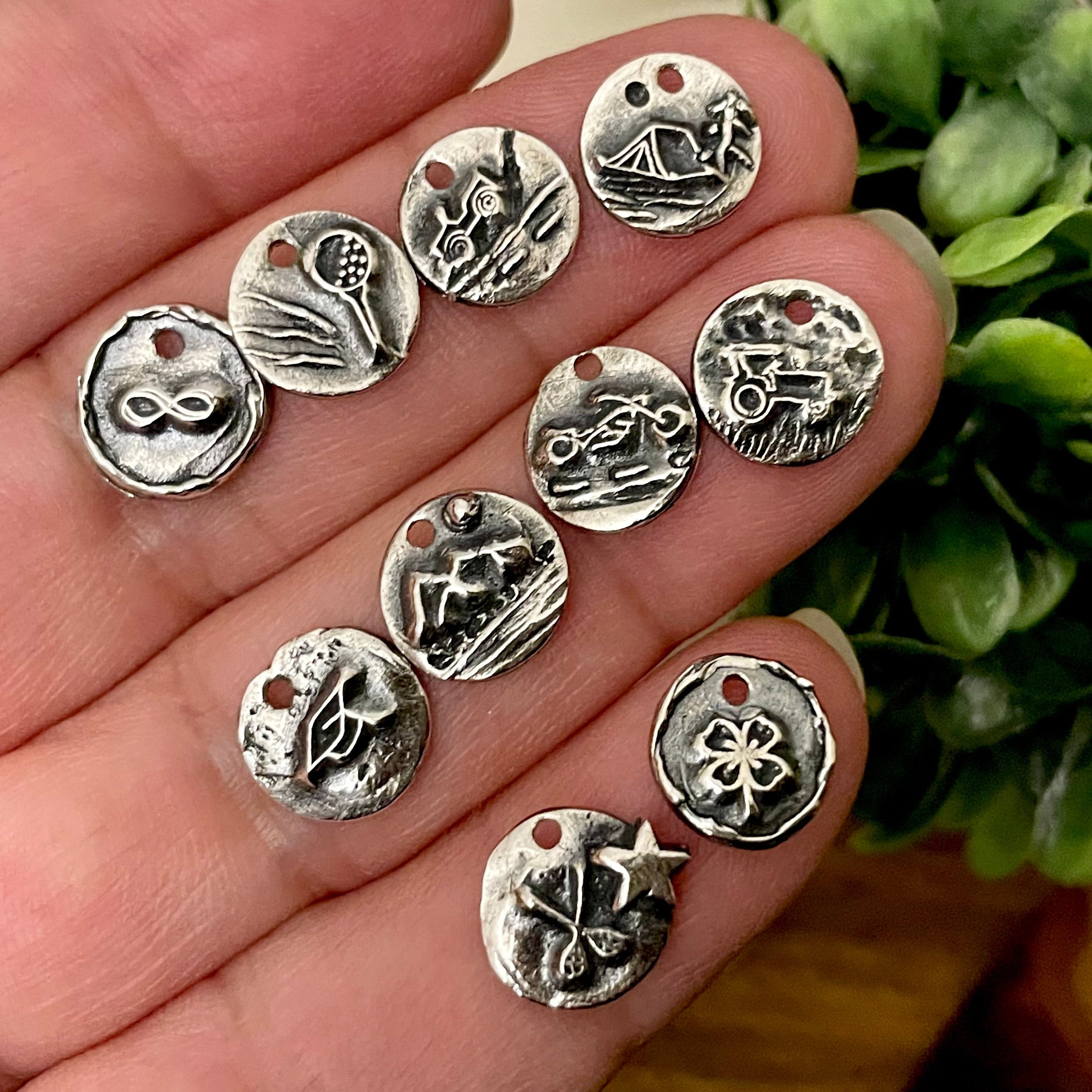 Sterling Silver charms with various designs held in a hand, with a green plant in the background.