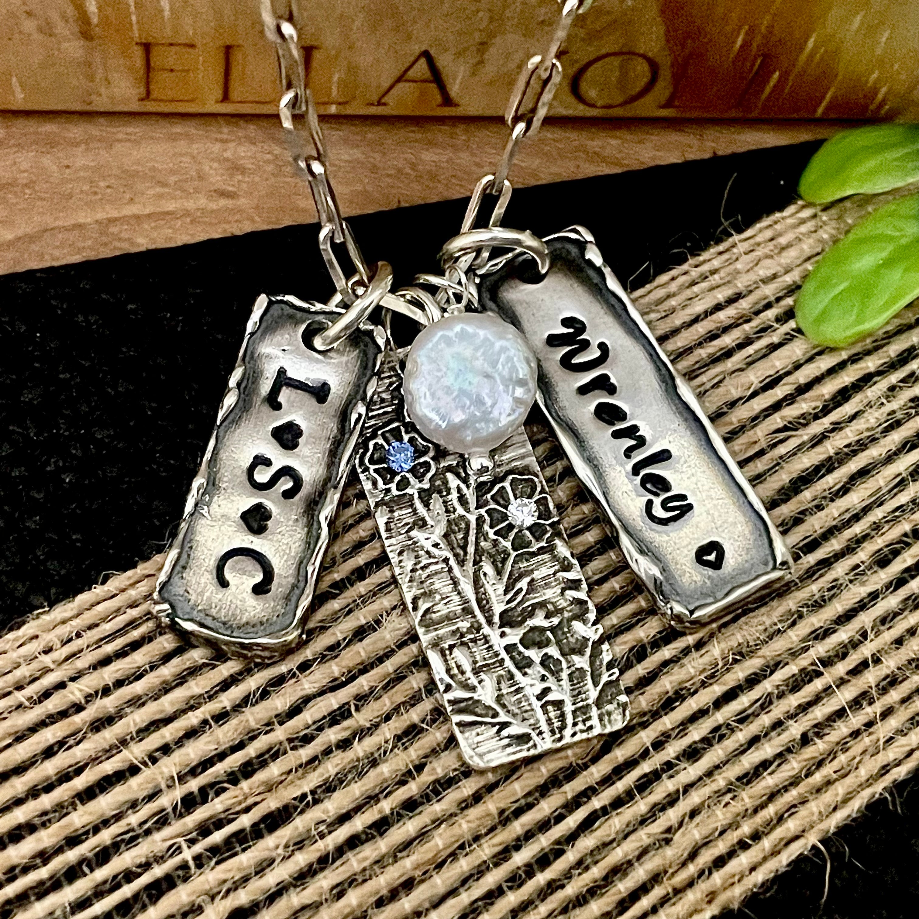 Silver necklace with engraved names, birthstones, initials, heart, flowers and pearl on sterling silver chain, displayed on a textured surface.