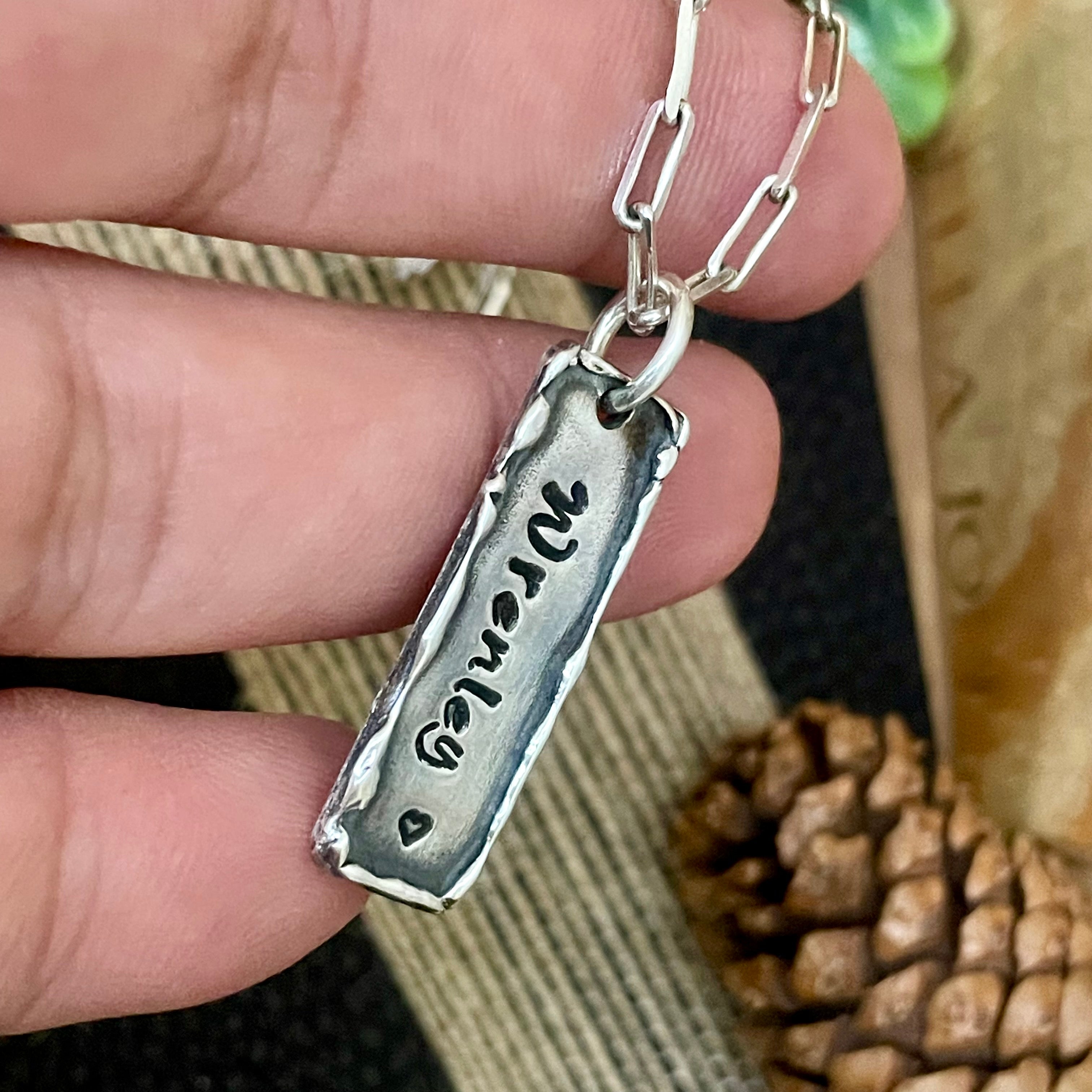 Silver necklace with a personalized rectangle organic boho style bar pendant held by a hand, with a blurred background. Custom pendant has engraved name and small heart below it. 