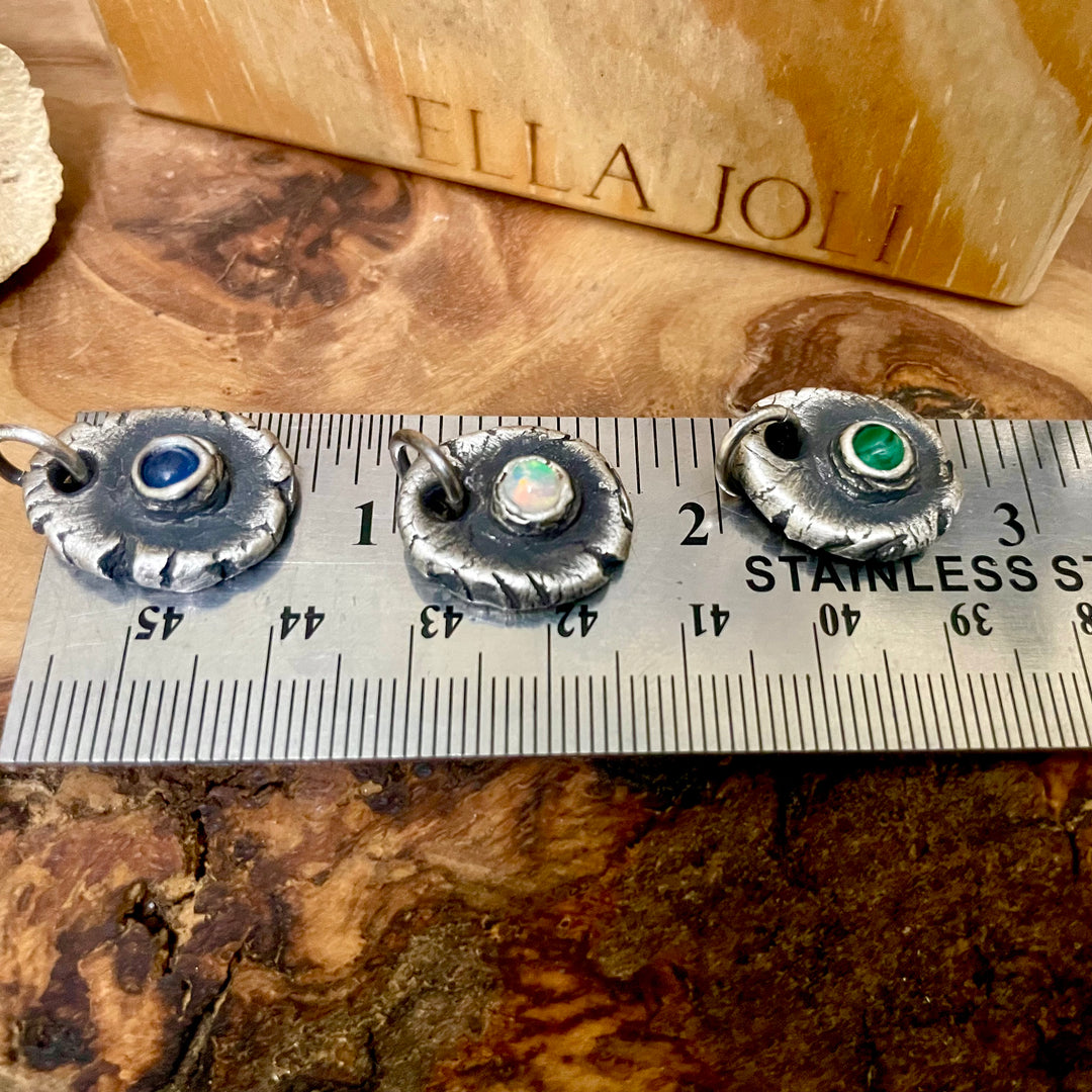 Three silver pendants with gemstones on a ruler for scale, against a wooden background with &