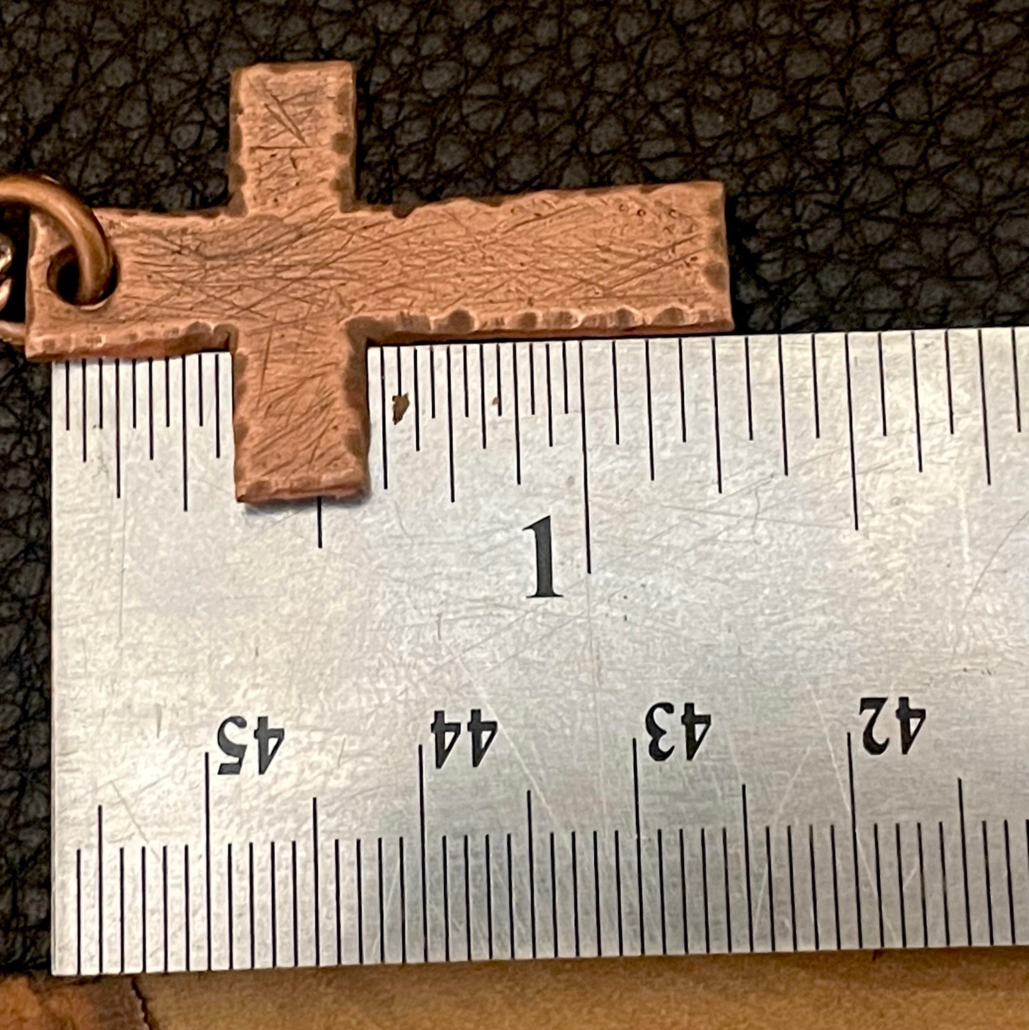 Copper cross pendant next to a ruler on a textured surface