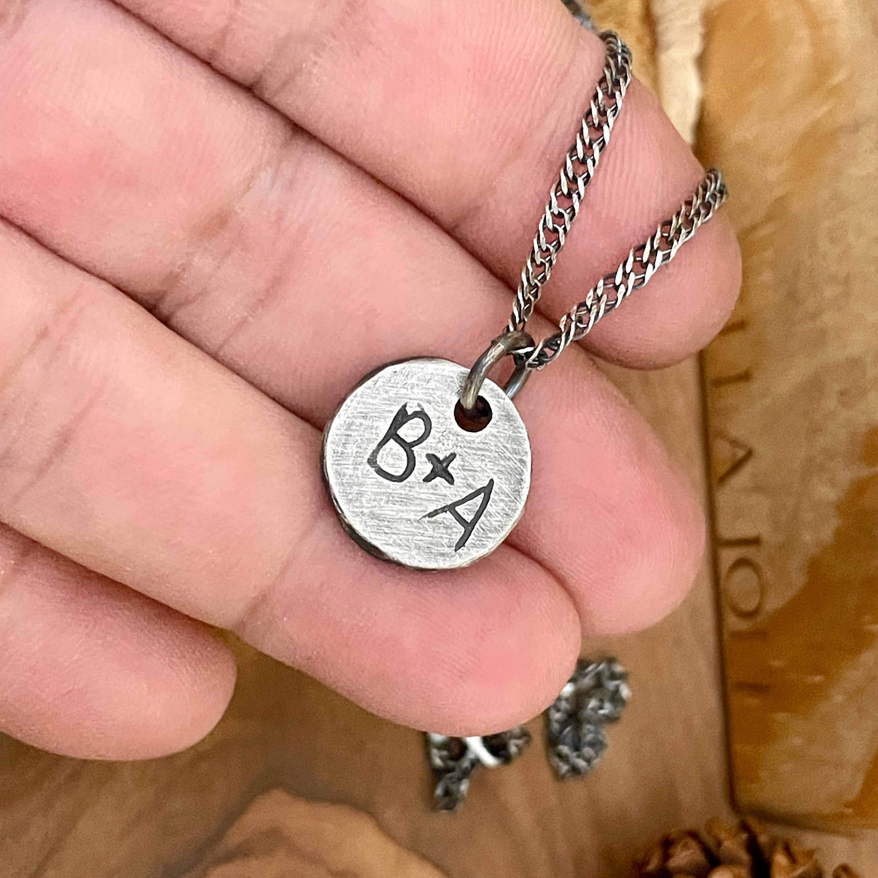 Silver necklace with a round pendant featuring 'BxA' held in a hand on a wooden surface.