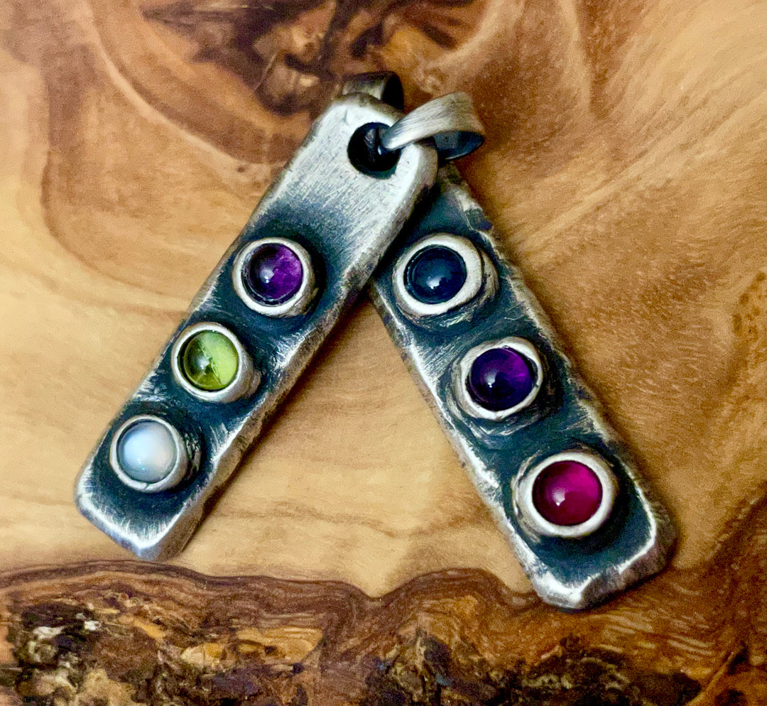 Two oxidized sterling silver pendants with three natural gemstone birthstones set into bezel settings. Handmade laying on wood display. Blue, Purple, Green, White, Pink shown. 