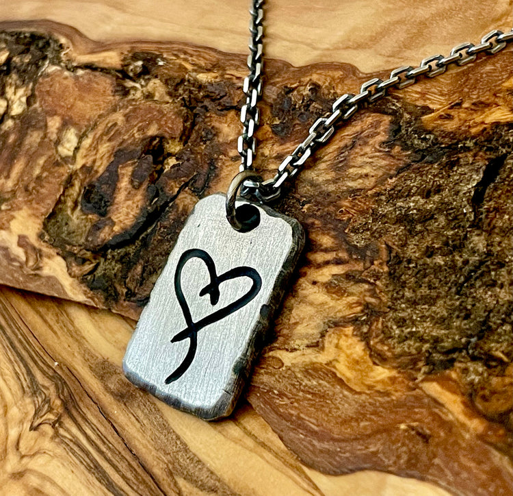 mens sterling silver necklace pendant hand carvbed with customers tattoo image into surface. Hanging from Cable chain on wood background  This example photo shows a heart. Hand forged by Ella Joli