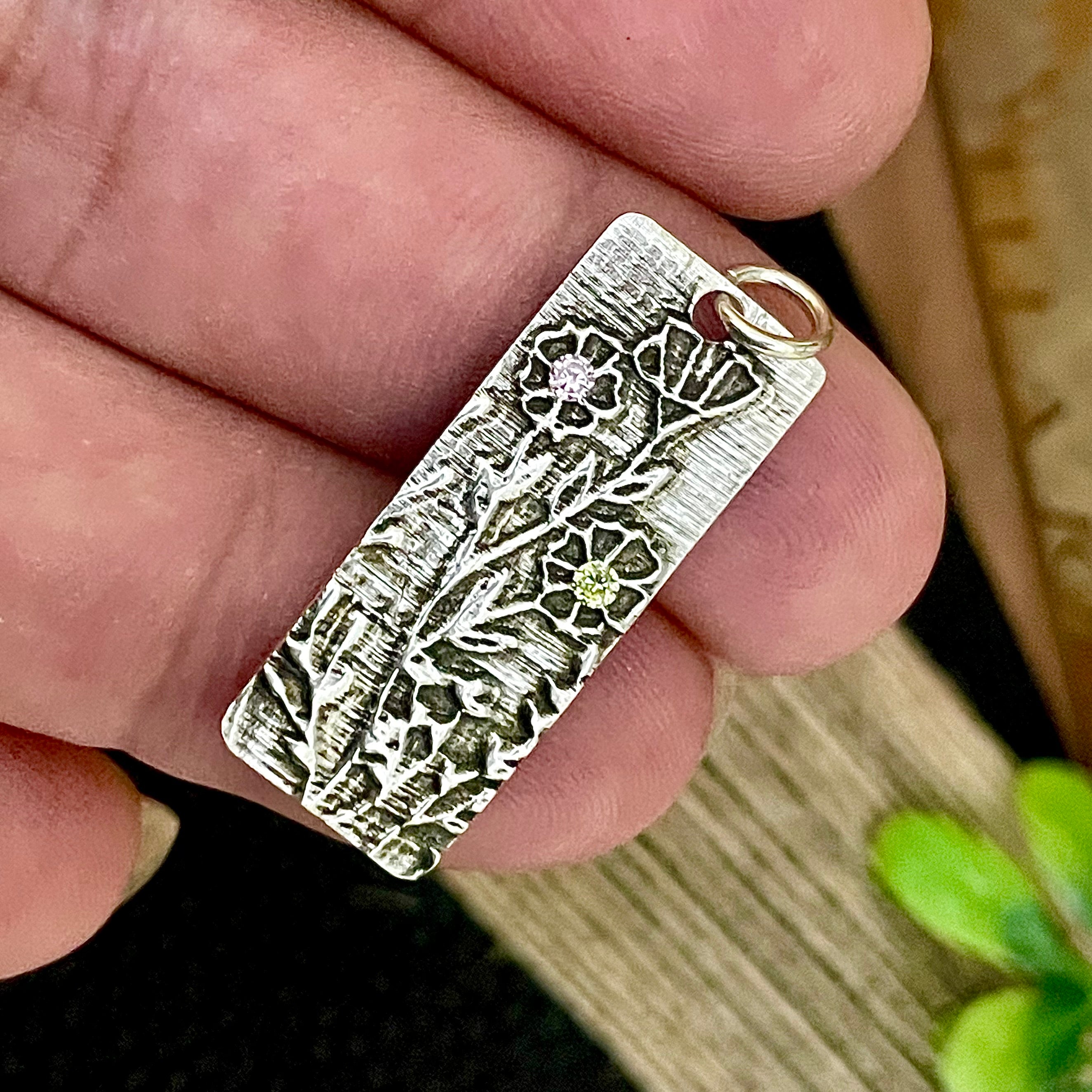 Silver pendant with intricate design held between fingers against a blurred background
