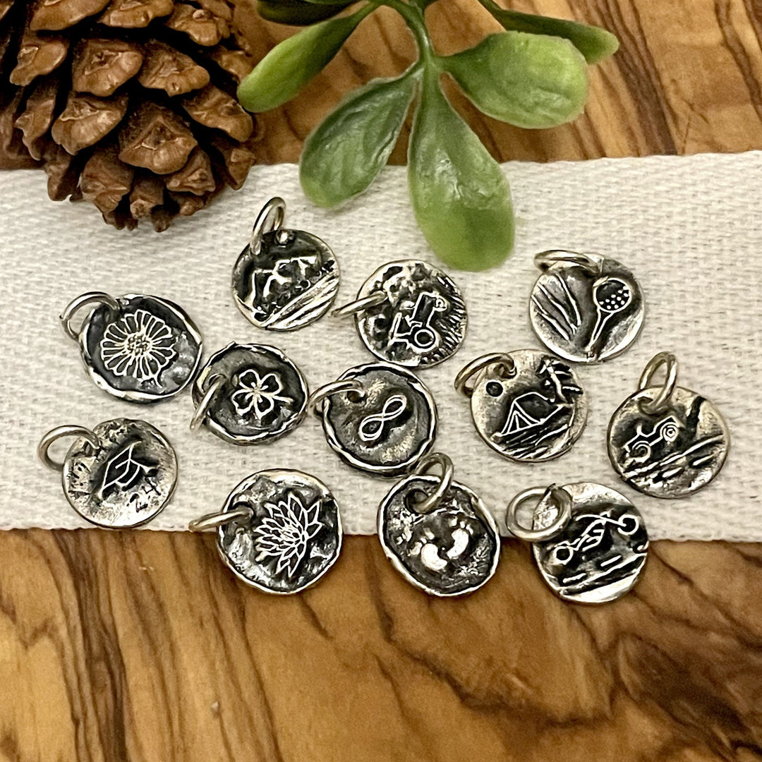 Collection of silver charms on a wooden surface with a pine cone and plant. Themes are outdoors, sports, family, friends, baby, travel, floral, farming, ranching, camping, graduation, luck, love, western, holiday, occasions, religion, birthday, Mothers day, minimal, boho, modern, personalized, custom made with intials on back. 
