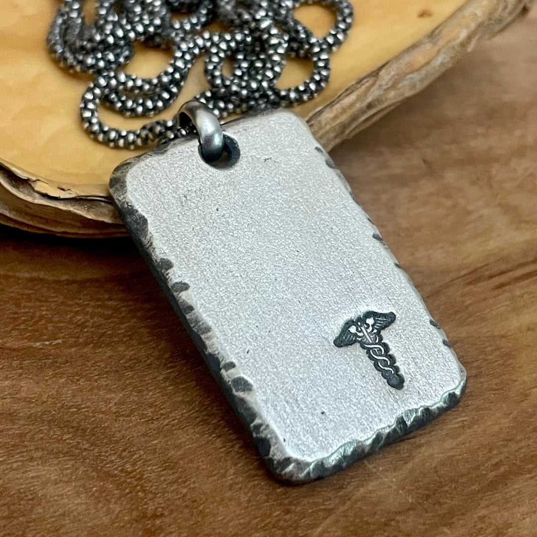 Oxidized sterling silver medical alert pendant with textured edges and symbol. Hanging from sterling silver chain laying on wood background. Choose 1 inch or 1.5 inch customized tag. Ella Joli