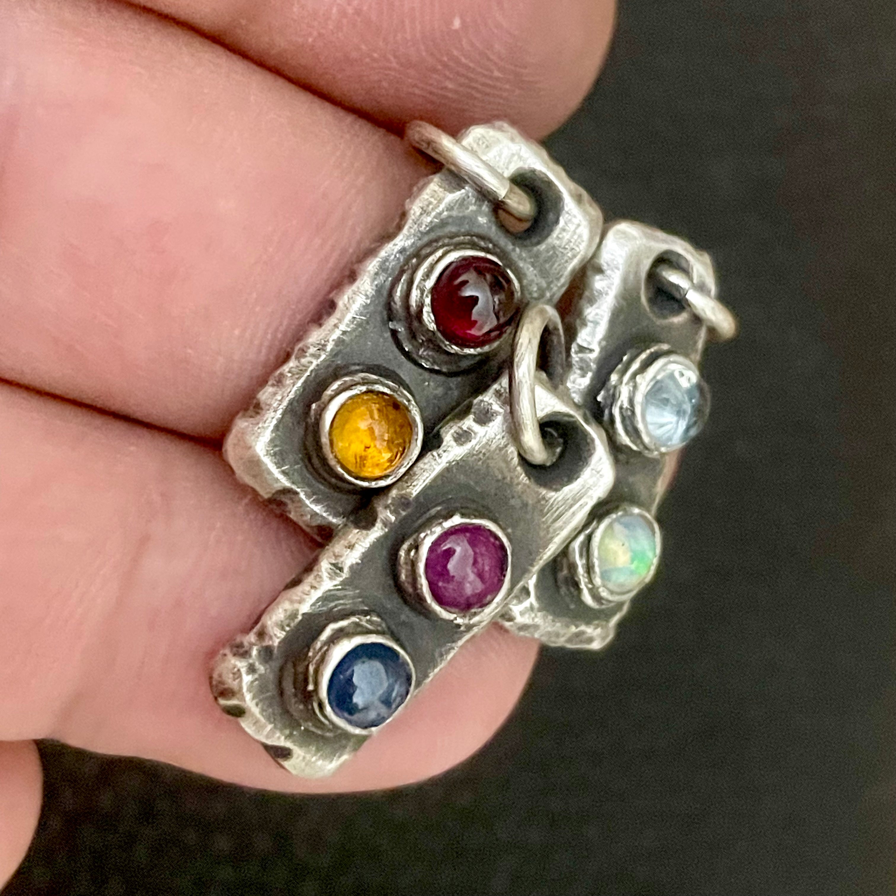 An assortment of oxidized Sterling Silver pendants with colorful birthstones/natural gemstones held in a hand against a dark background. Each rectangle bar pendant/charm has two birthstones of choice. Family Jewelry Gift for Him. 