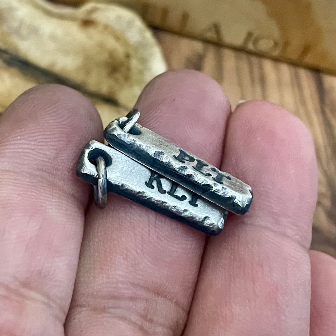 Two silver pendants on a finger with 'PLT' and 'KLI' engraved.