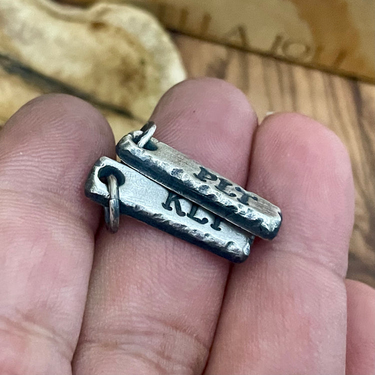 Two silver pendants on a finger with 'PLT' and 'KLI' engraved.