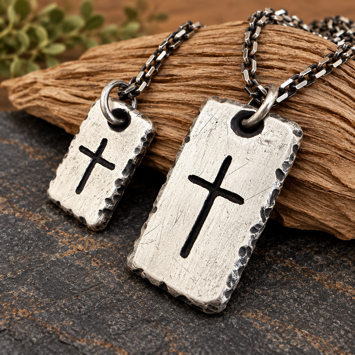 Two solid fine silver rectangular pendant necklaces hand forged with  engraved crosses, displayed on a piece of wood over a dark textured surface.