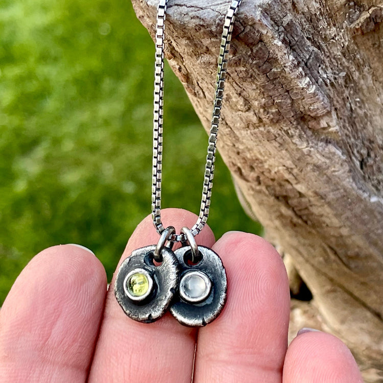 Two birthstone pendants hanging from oxidized sterling silver box chain in hand with nature background