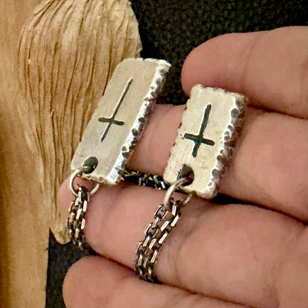 A close-up of a hand holding a pair of silver-toned rectangular drop earrings with engraved cross symbols and textured edges, shown against a dark textured background with a wooden surface on the left.