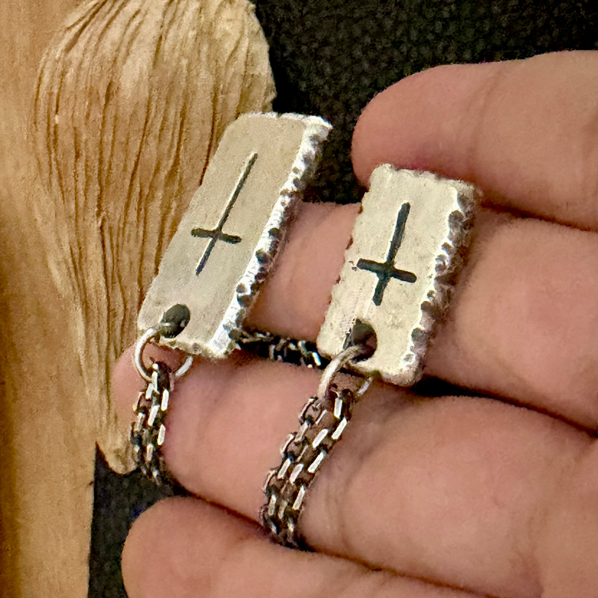 A close-up of a hand holding a pair of silver-toned rectangular drop earrings with engraved cross symbols and textured edges, shown against a dark textured background with a wooden surface on the left.