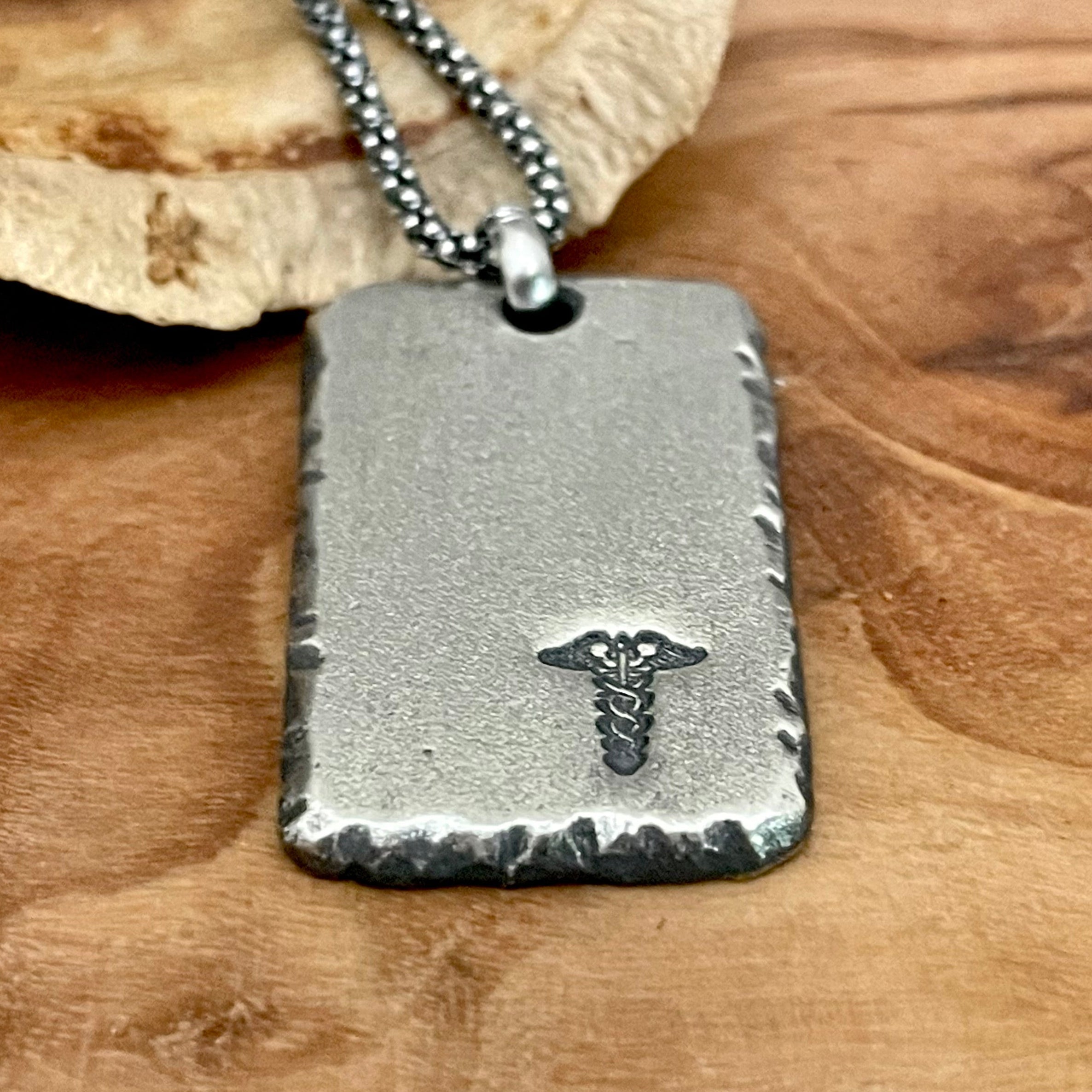 thick sterling silver medical alert pendant tag that can be engraved on back side. Hanging from sterling silver chain laying on wood back ground
