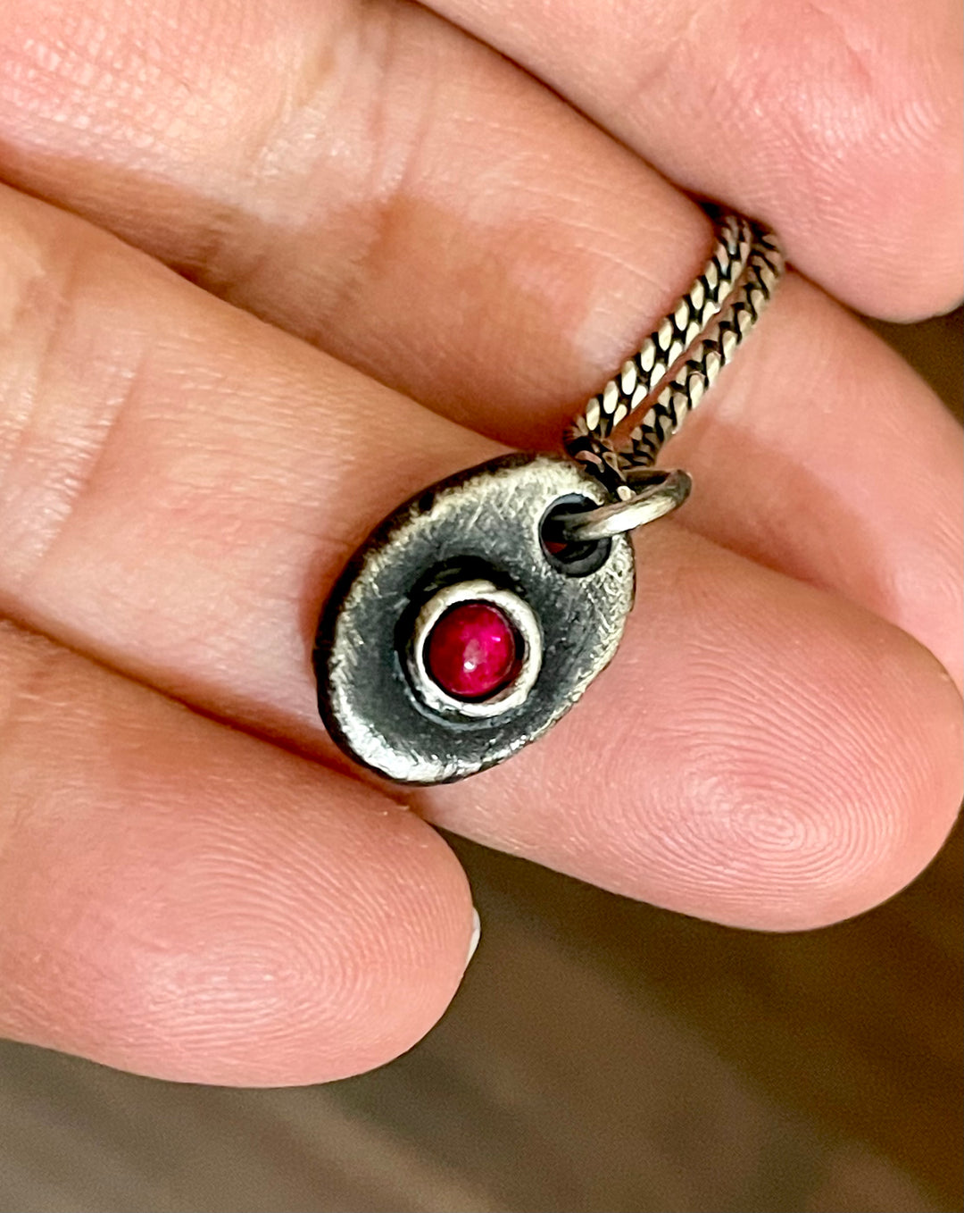 men's ruby necklace and pendant in hand. Made from oxidized sterling silver in oval shape with round stone.