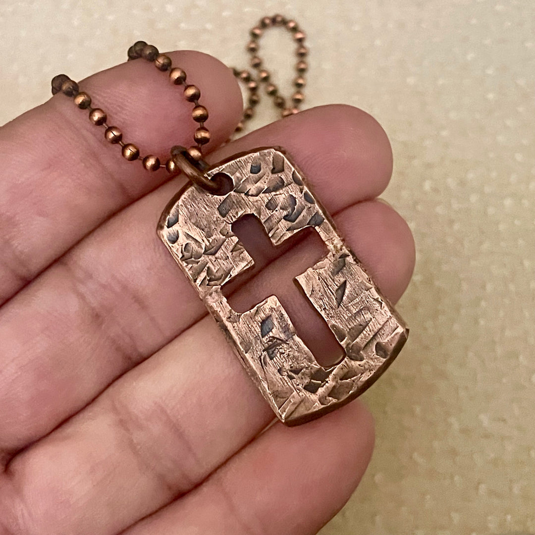 antique copper cross pendant held between fingers against a beige background hanging from copper ball bead chain
