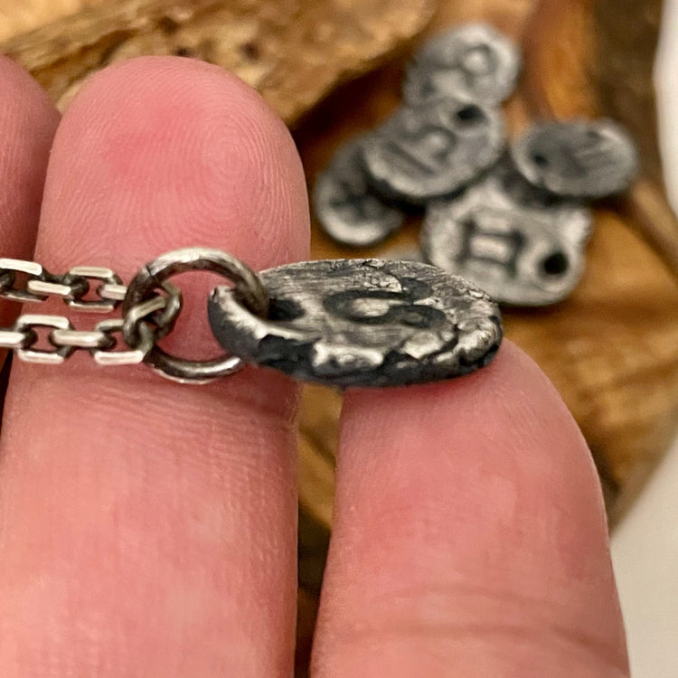 Side view of thick and textured oval to round shaped zodiac symbol necklace hanging from ring on 2.7mm cable chain. Shown in hand with other charms in background on wood surface. 
