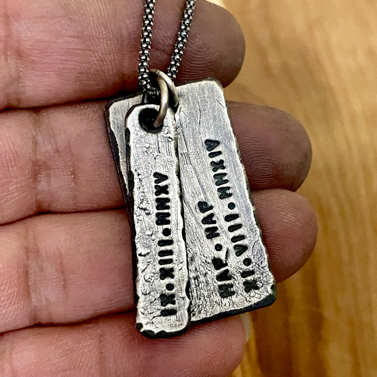 Handcrafted rustic silver pendant necklace on hand with wood background. Roman numerals and initials are engraved on pendants hanging from oxidized silver chain. 