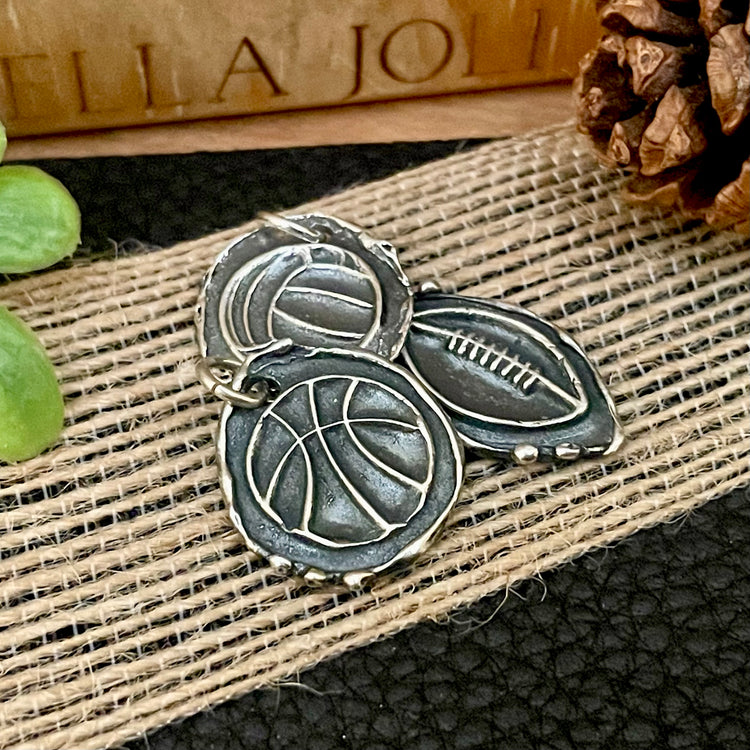 Three oxidized sterling silver sports ball pendants or charms that can be personalized on backside. Sitting on a textured surface and pine cone in the background. Basketball, football and volleyball shown with baseball/softball and soccer also available. 