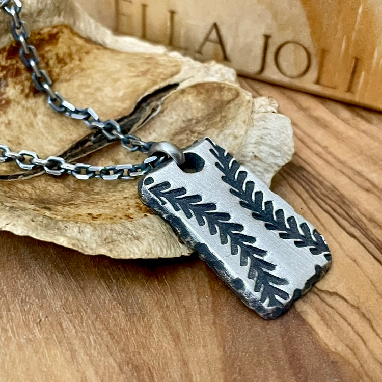 One inch rectangle shaped baseball pendant with hand carved ball stitches. Oxidized to darken recessed areas. Hanging from 2.7mm sterling silver chain, displayed on wood background with designer Ella Joli text in background. 