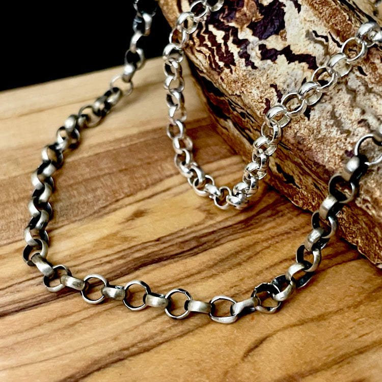 A 4mm sterling silver rolo chain necklace with a matte oxidized finish, and a polished finish, displayed on a wooden surface.