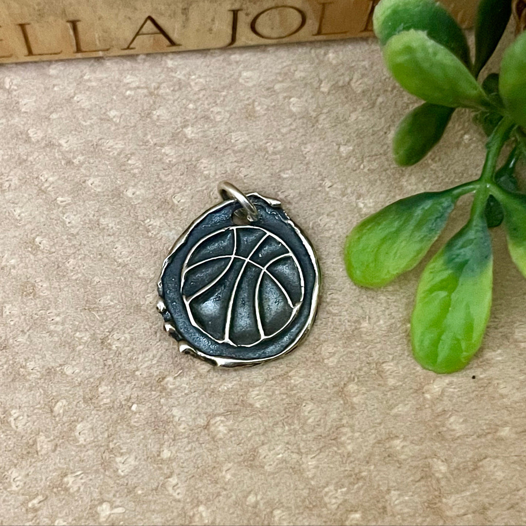 oxidized sterling silver basketball charm with raised artisan details. Can be personalized with name or number on backside. Displayed on cream and wood background with green accent. 