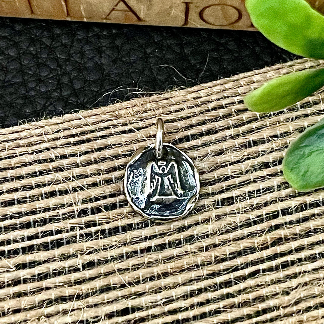 Sterling silver angel small pendant or charm with raised and engraved design on a textured fabric background. Handmade Artisan Silver pendant on a textured surface with a plant leaf in the corner