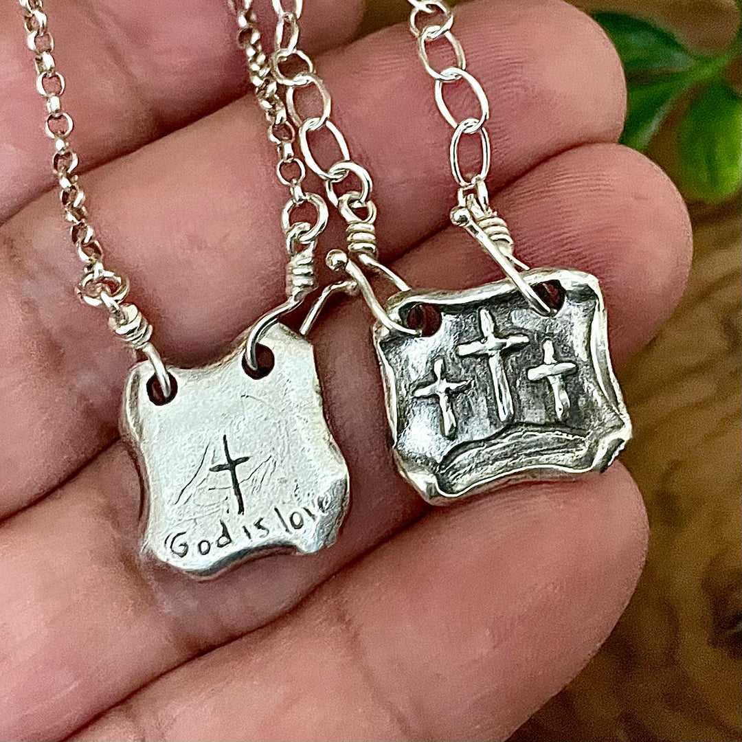 Two sterling silver pendants with raised design of three crosses on front of pendant and "God is love" shown hand carved onto back side of pendant with cross. Both hanging from sterling silver interchangeable chains, being held in a hand.
