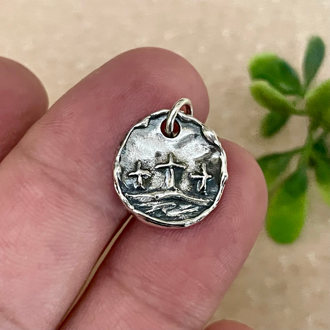 Silver pendant with raised design of three crosses held between fingers against a blurred natural background
