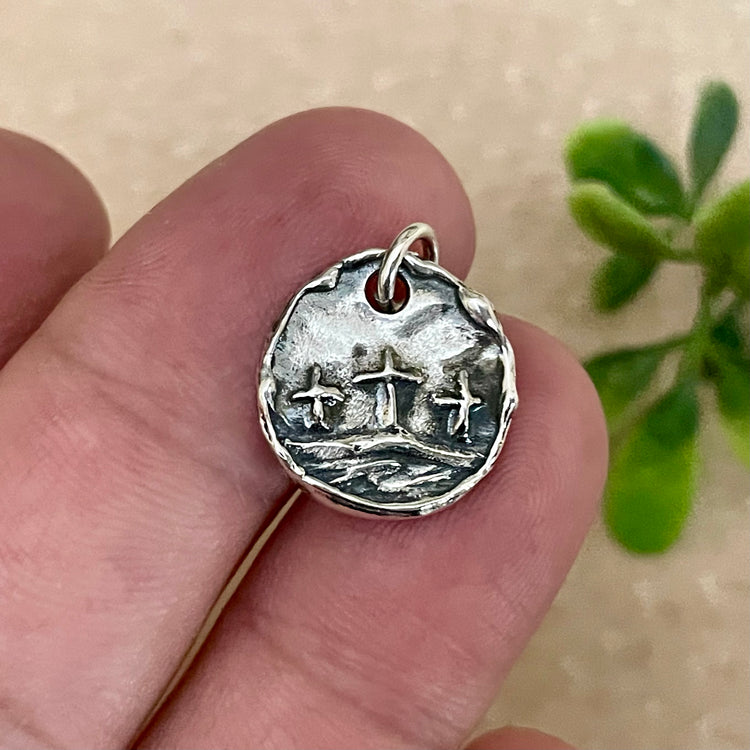 Silver pendant with raised design of three crosses held between fingers against a blurred natural background