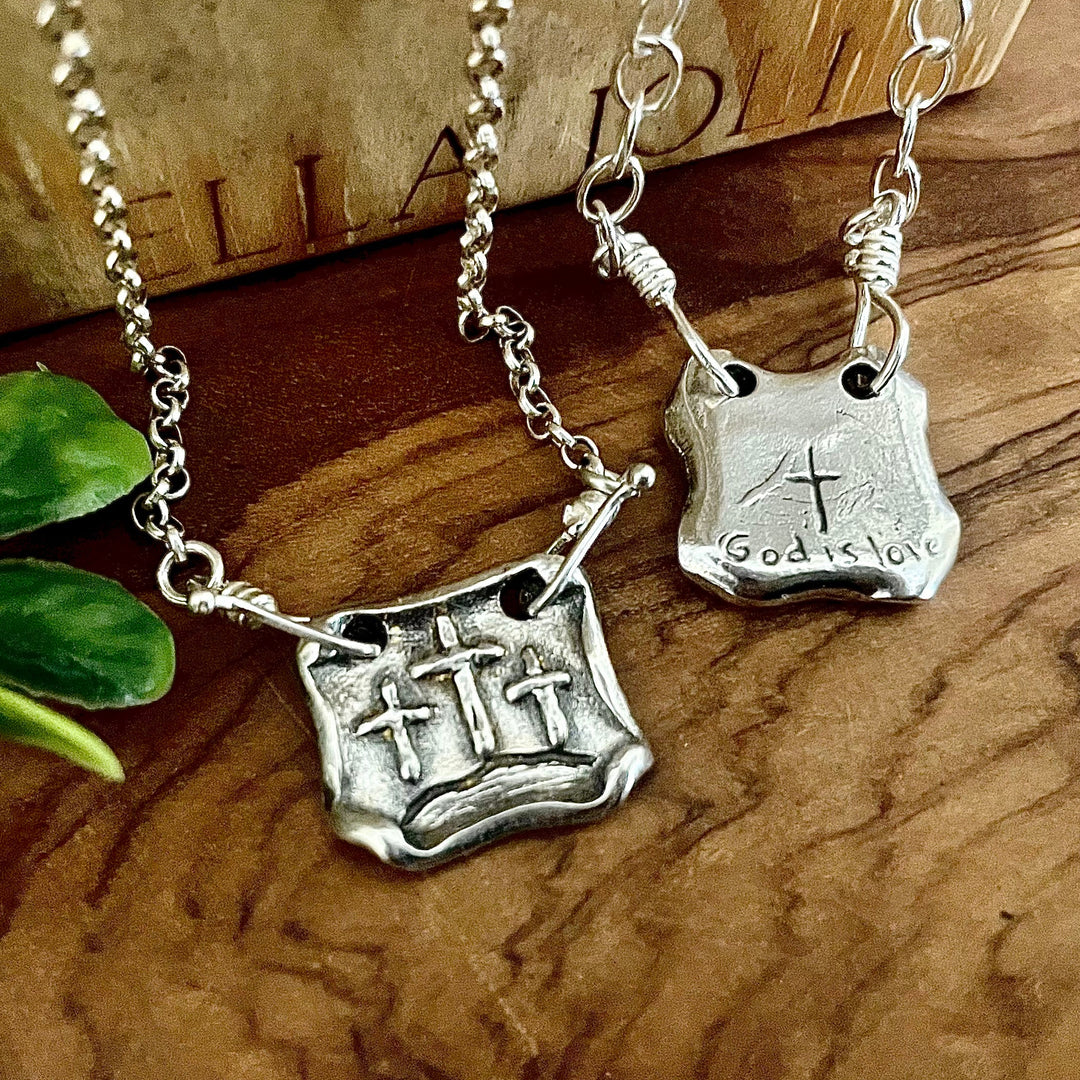 Artisan handmade square shaped pendant with scroll edges and three raised cross images to represent the three crosses of calvary. Backside of second silver pendants shows hand carved God is love words with a cross. Displayed on wooden background. 