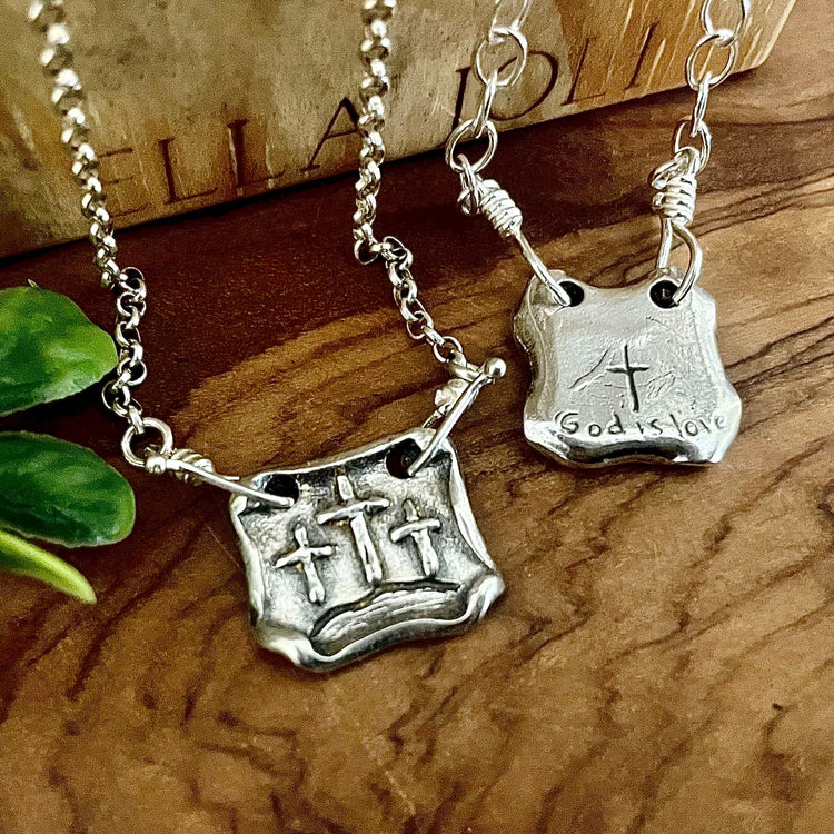 Artisan handmade square shaped pendant with scroll edges and three raised cross images to represent the three crosses of calvary. Backside of second silver pendants shows hand carved God is love words with a cross. Displayed on wooden background. 