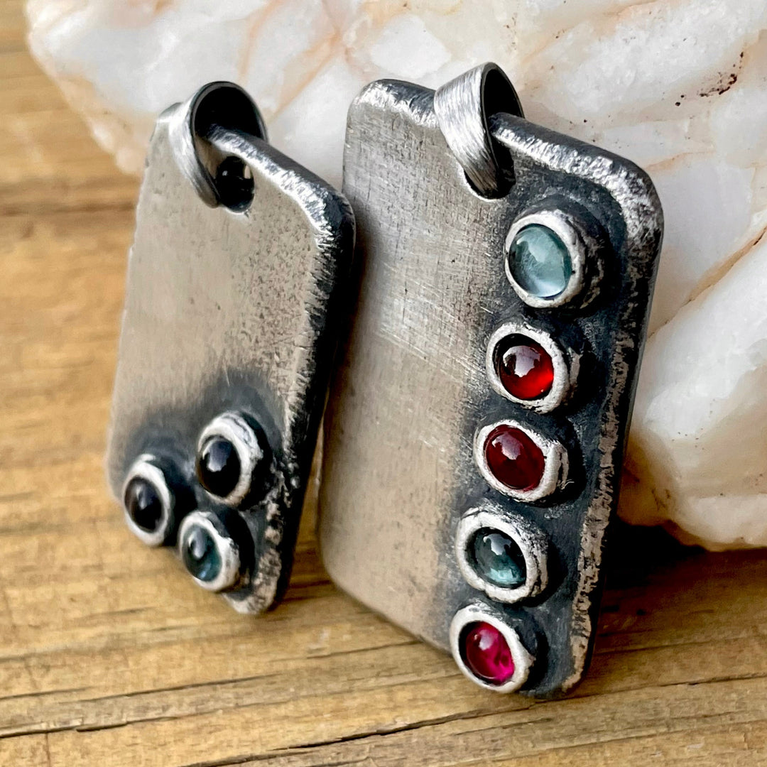 Two rectangular silver birthstone pendants with multiple colored gemstones set in them, displayed on a wooden surface.