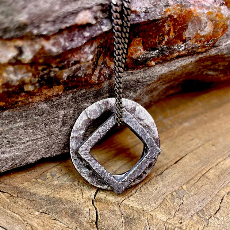 Hand carved pendants stacked together hanging from 2mm curb chain. Bottom pendant is an oxidized circle and top pendant is an oxidized square pendant. Free floating charms on 2mm curb chain. Shown on wood and stone background. 