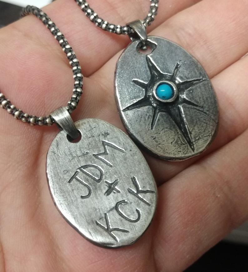 Back side of sterling silver compass necklace with turquoise gemstone showing hand carved initials on back and turquoise stone on front. Laying in hand. 