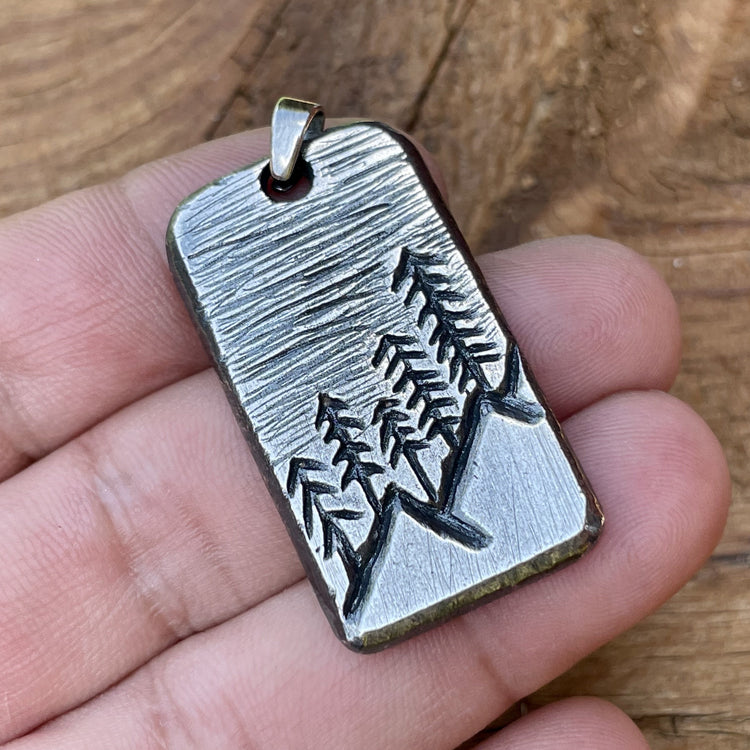 Oxidized sterling silver hand carved rectangle pendant with mountain and tree scene. Back side can be personalized. Shown in hand.  