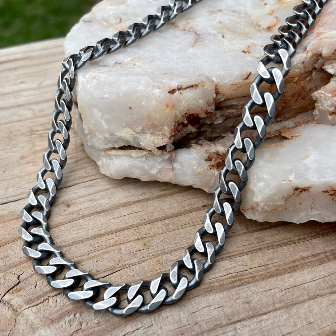 Heavy oxidized sterling silver curb chain necklace 10mm in width with lobster clasp, displayed on stone and wood. 